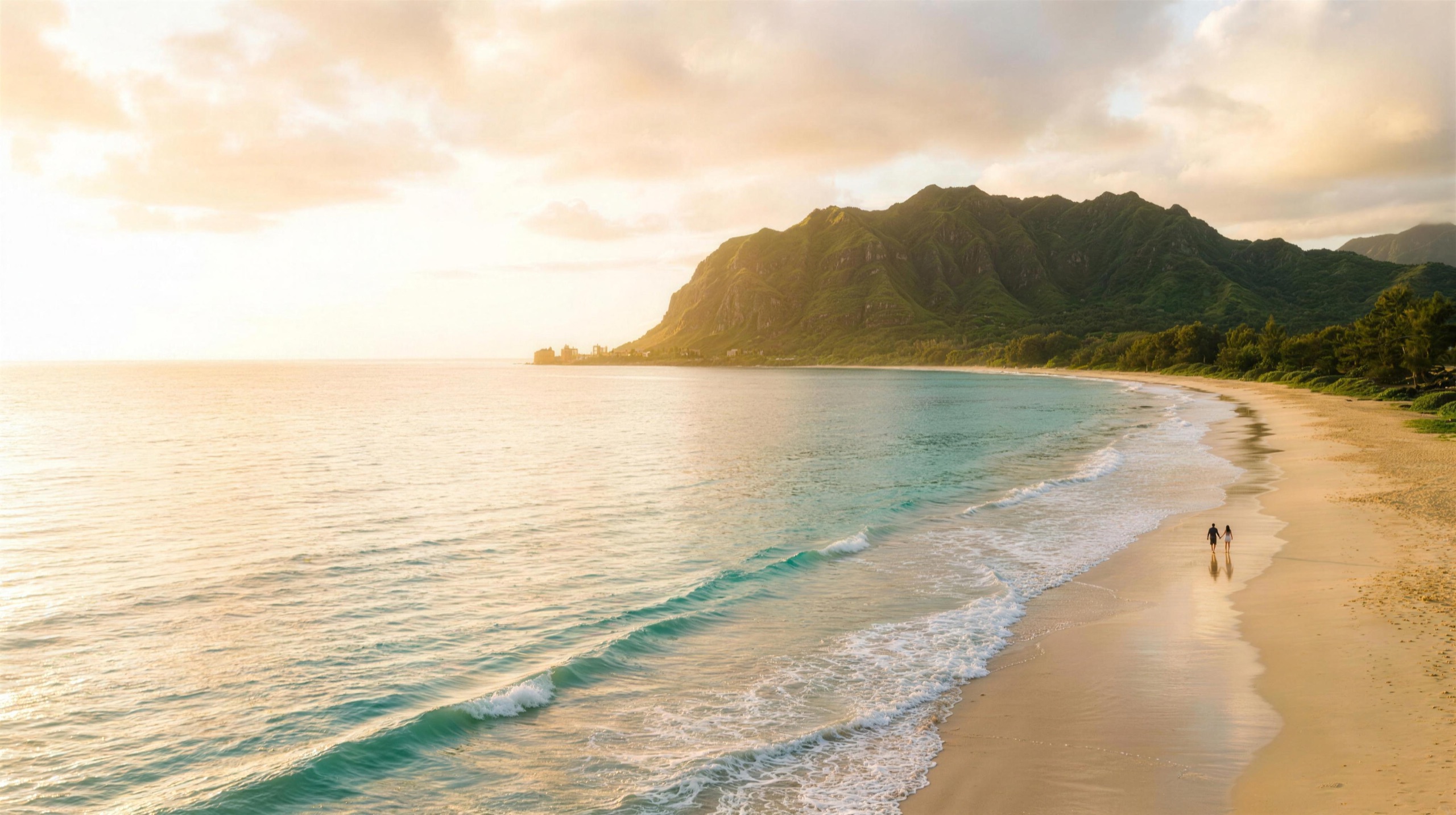 Golden-hour aerial of an Oʻahu beach - turquoise water, lush windward ridge, and a couple walking hand-in-hand along the shore